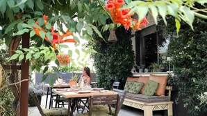 Un patio al aire libre con una mesa de comedor para dos personas, rodeado de abundante follaje y plantas en flor.