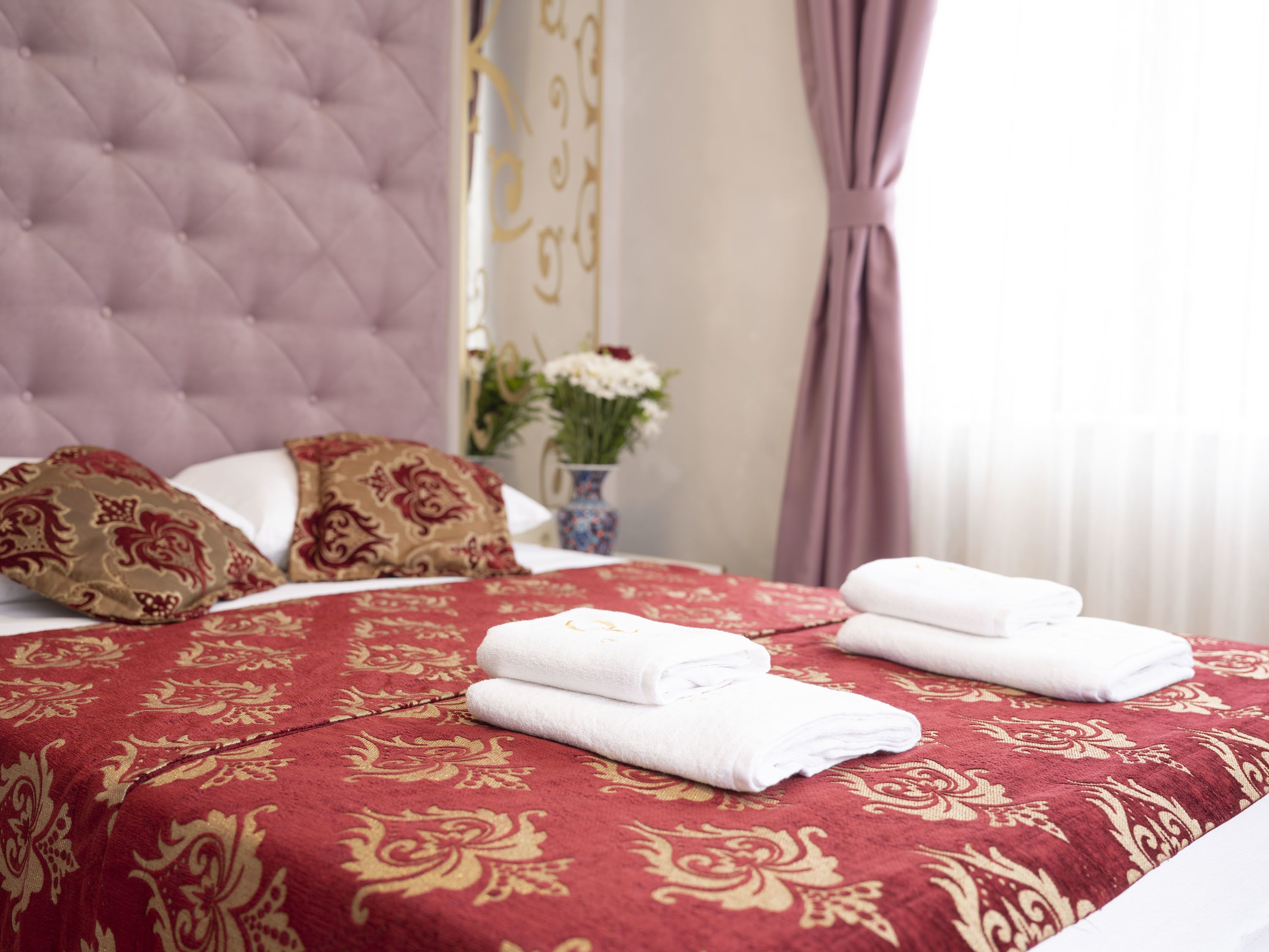 A neatly made bed with a red and gold patterned bedspread, two pillows with a red and gold design, and folded white towels on the bed.