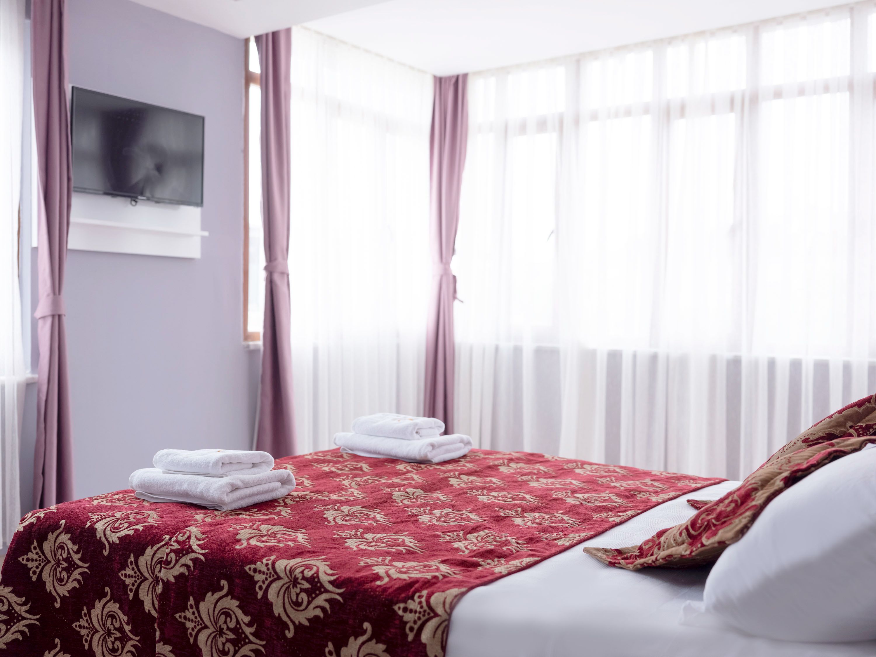 A hotel room with a neatly made bed, a television mounted on the wall, and a large window with sheer curtains.