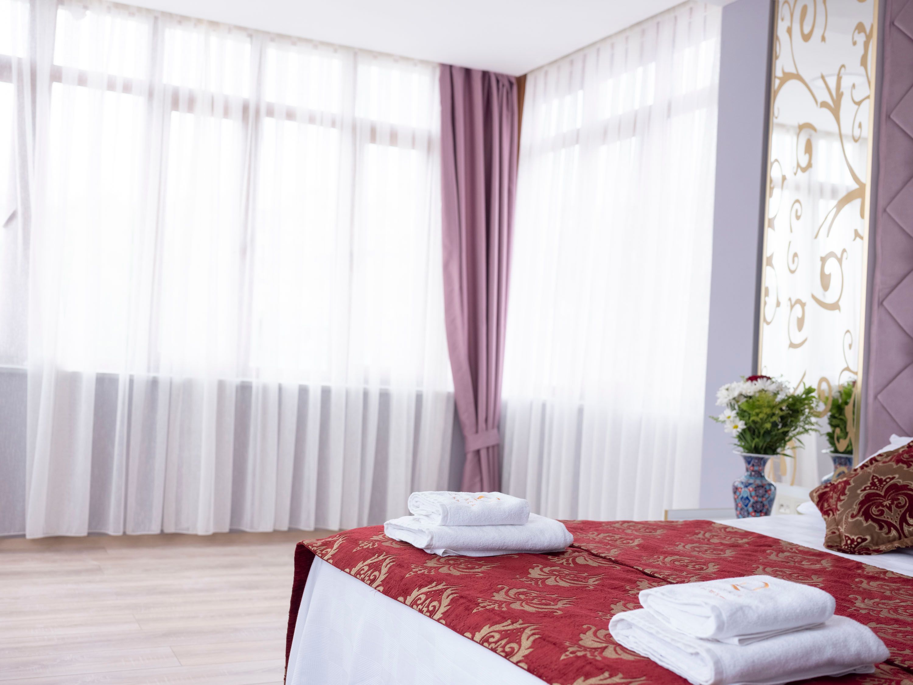 Habitación de hotel con cama, ventana con cortinas traslúcidas, colcha de diseño rojo y dorado, y un jarrón con flores.