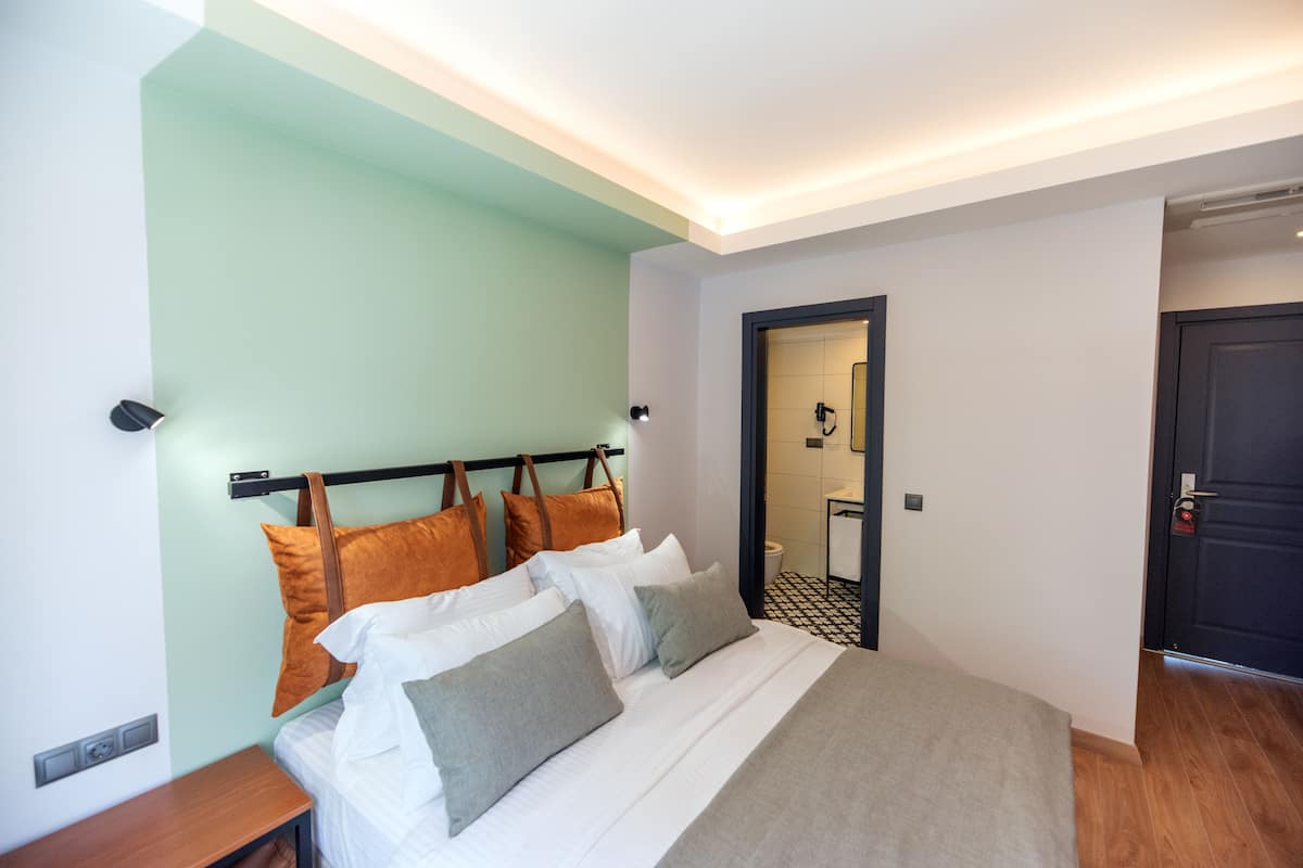 A modern hotel room with a neatly made bed, a bedside table, and a door leading to a bathroom.