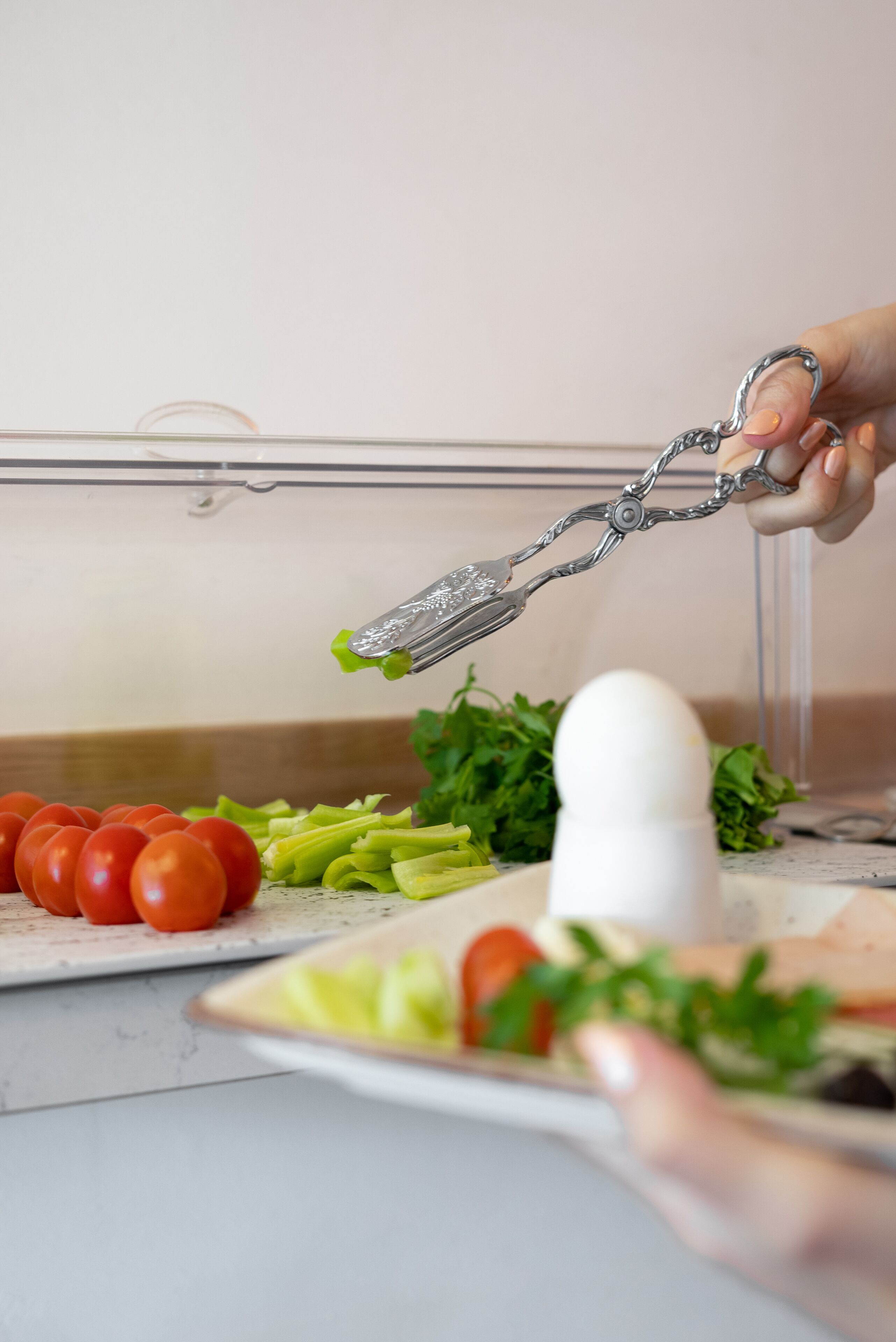 Une main tenant une pince au-dessus d’une assiette contenant des tomates, de la laitue et un œuf dur.