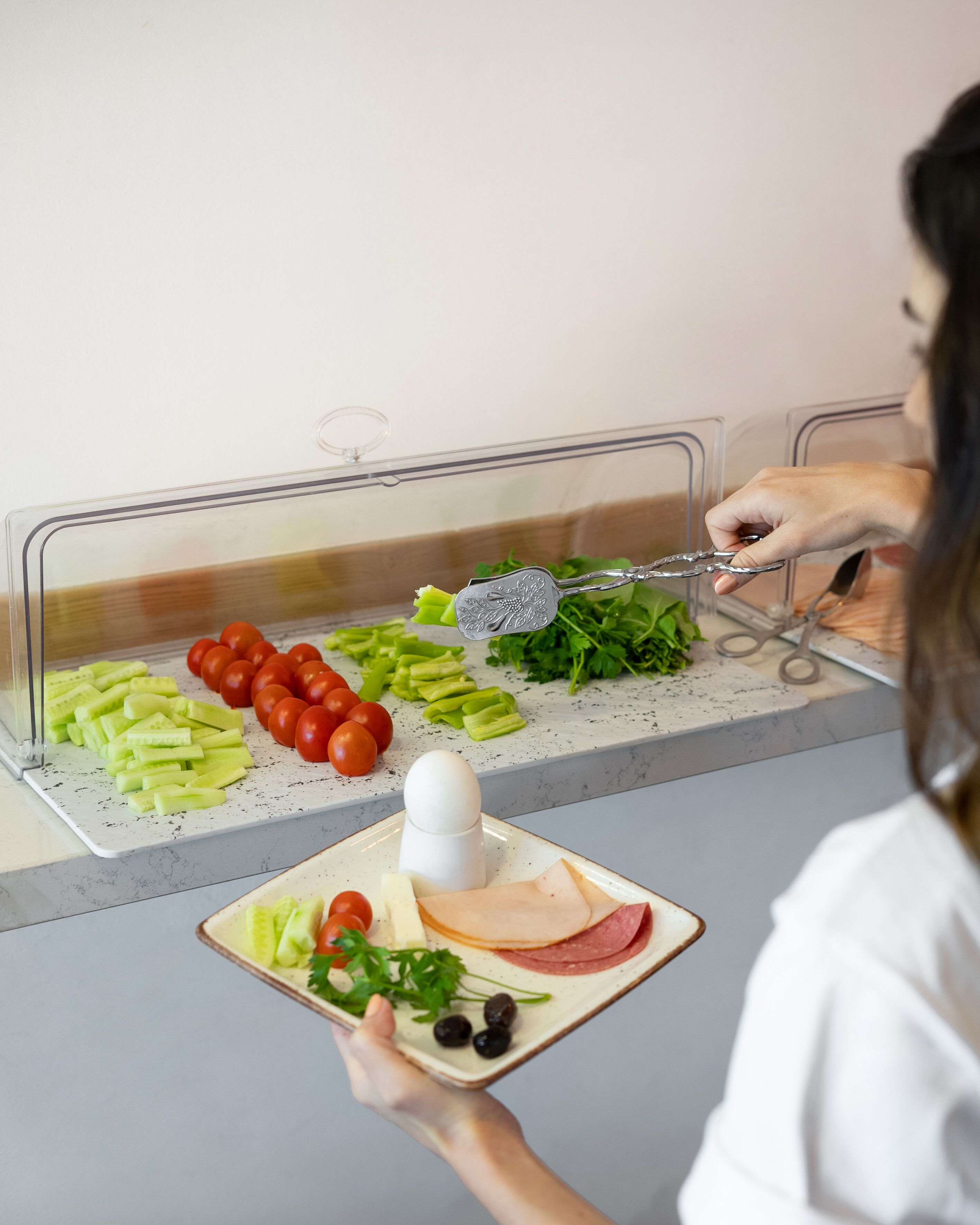 Una persona sosté un plat amb cogombre, tomàquets i olives, i utilitza pinces per agafar un tros de carn d'un mostrador de menjar.