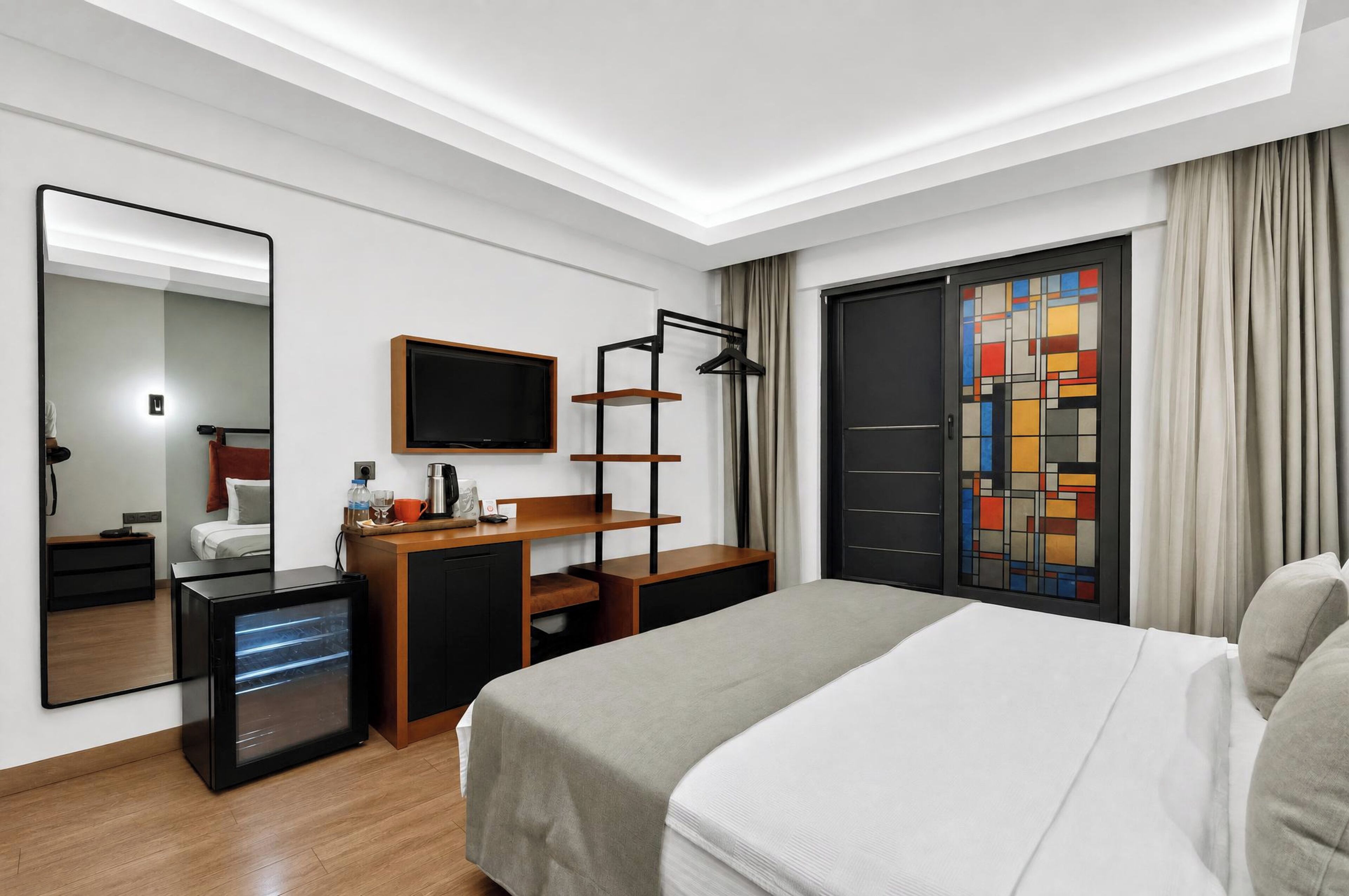 A modern hotel room with a bed, a desk with a computer, a TV, and a large window with curtains.