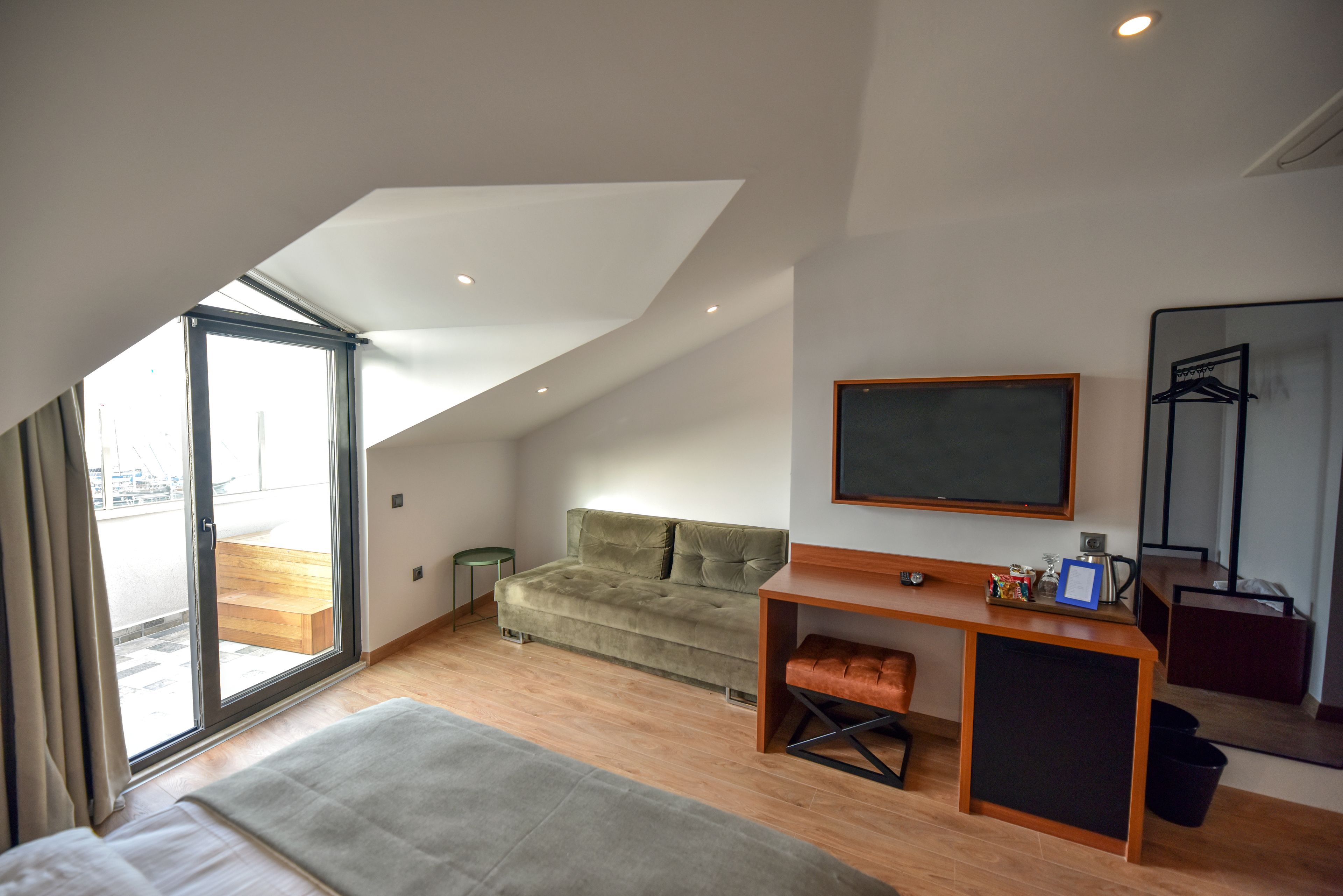 A modern hotel room with a large bed, a sofa, a desk with a flat-screen TV, and a sliding glass door leading to a balcony.
