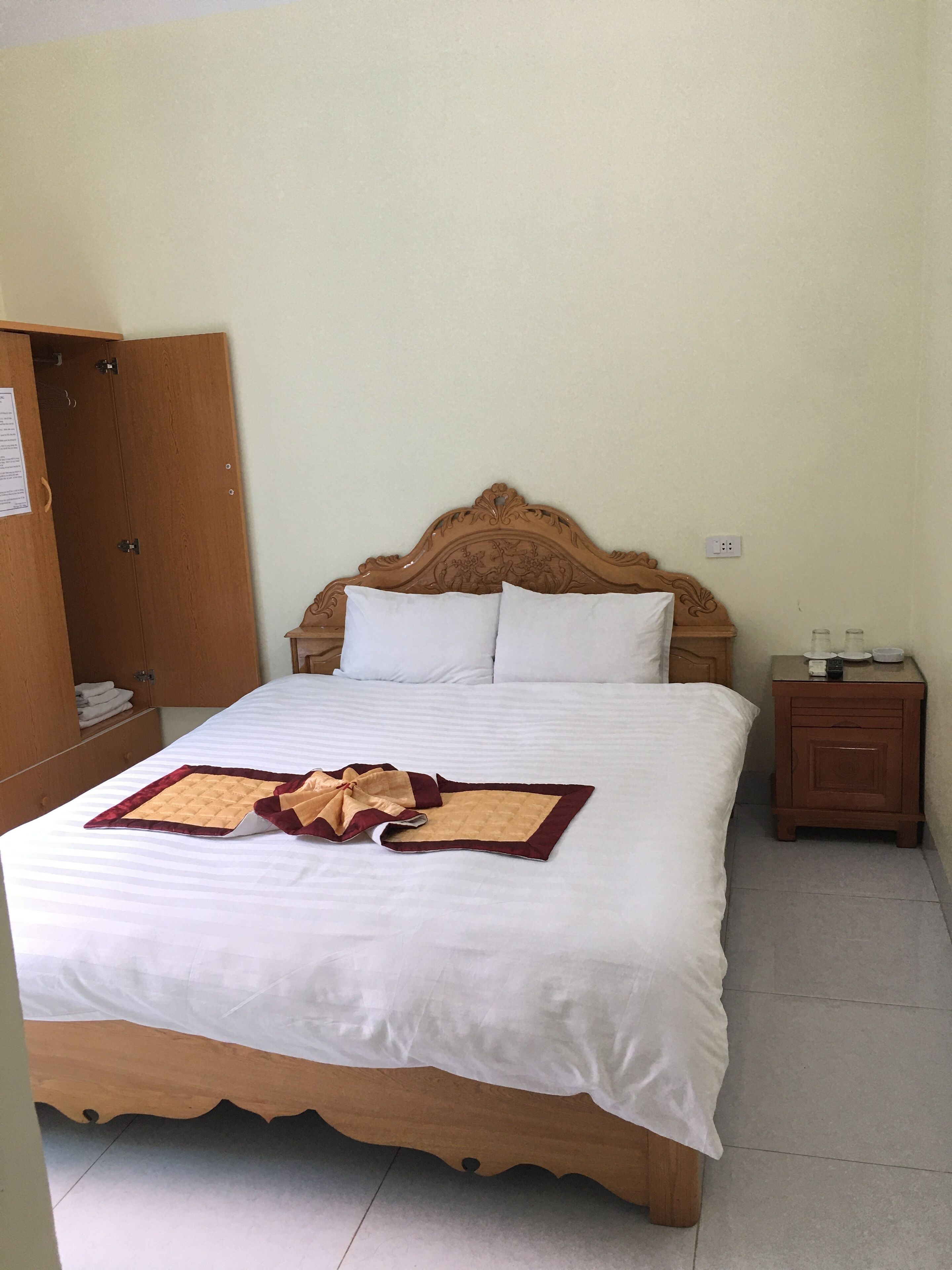 A neatly made bed with a white comforter and a decorative throw. A wooden wardrobe is to the left, and a small wooden nightstand with a lamp is to the right.