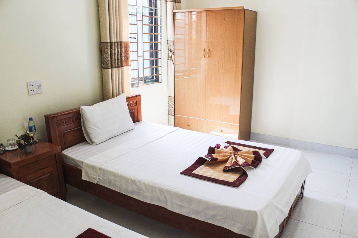 A hotel room with a bed, a wooden wardrobe, a nightstand, and a window with bars.