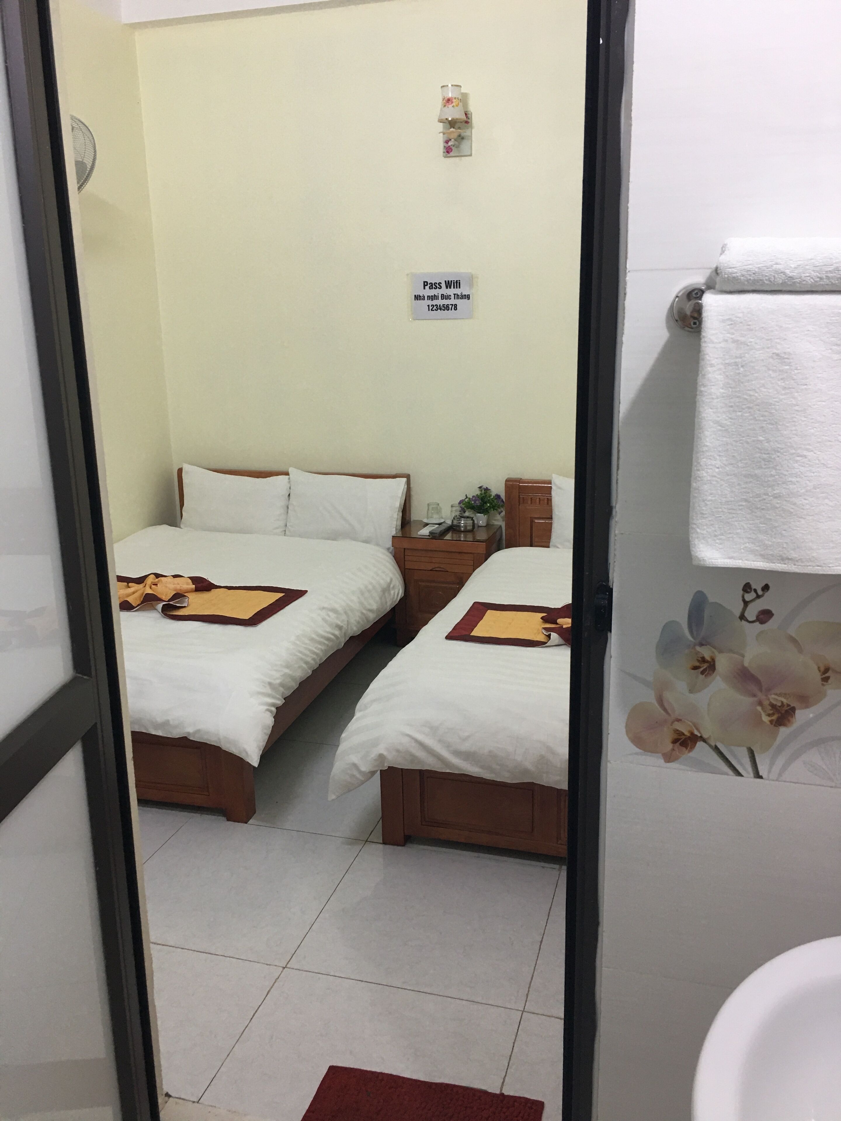 A hotel room with two beds, a small table, a nightstand, a sign indicating Wi-Fi availability, and a bathroom visible through an open door.
