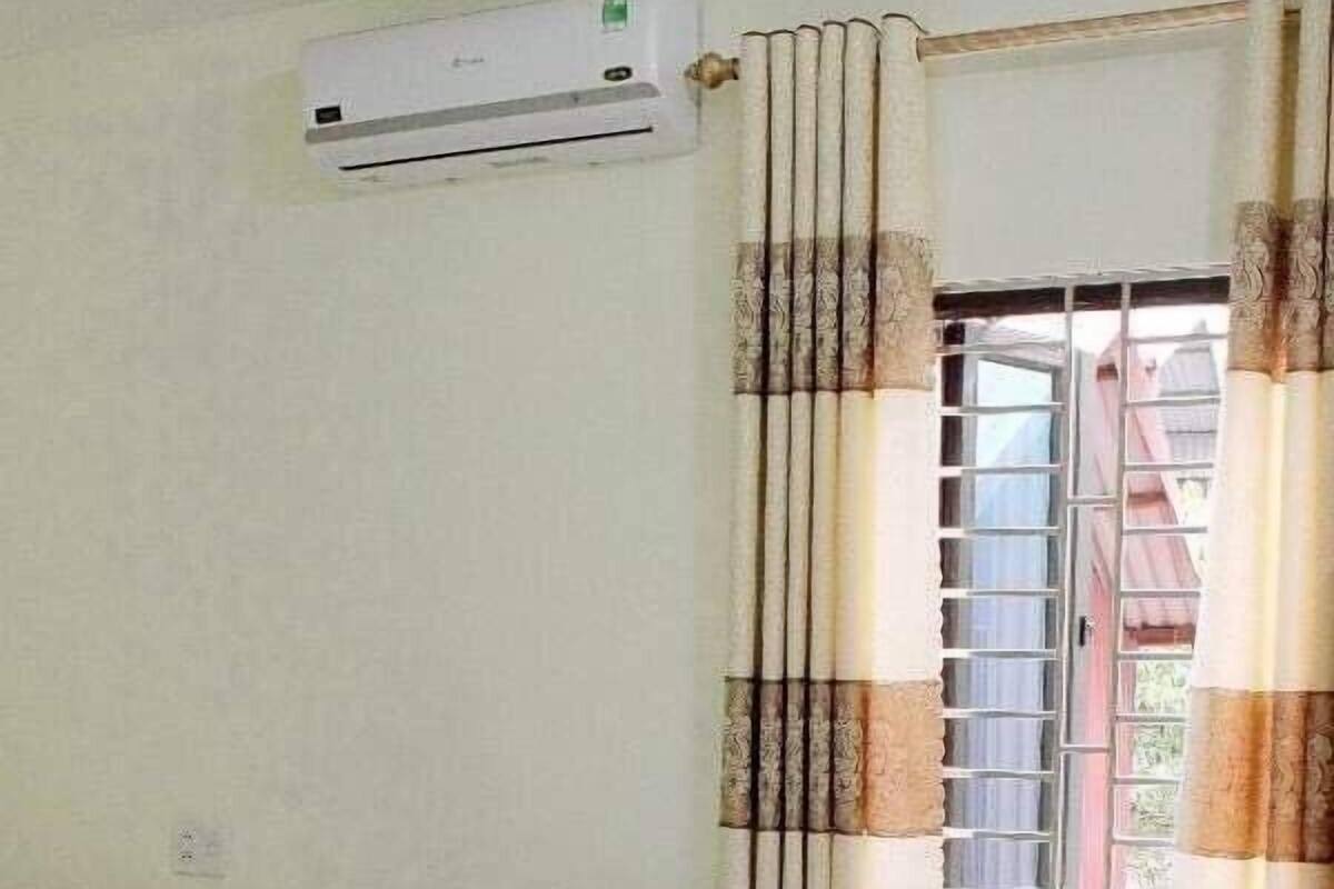 A hotel room with two beds, a window with curtains, an air conditioning unit, and a bedside table with a lamp.