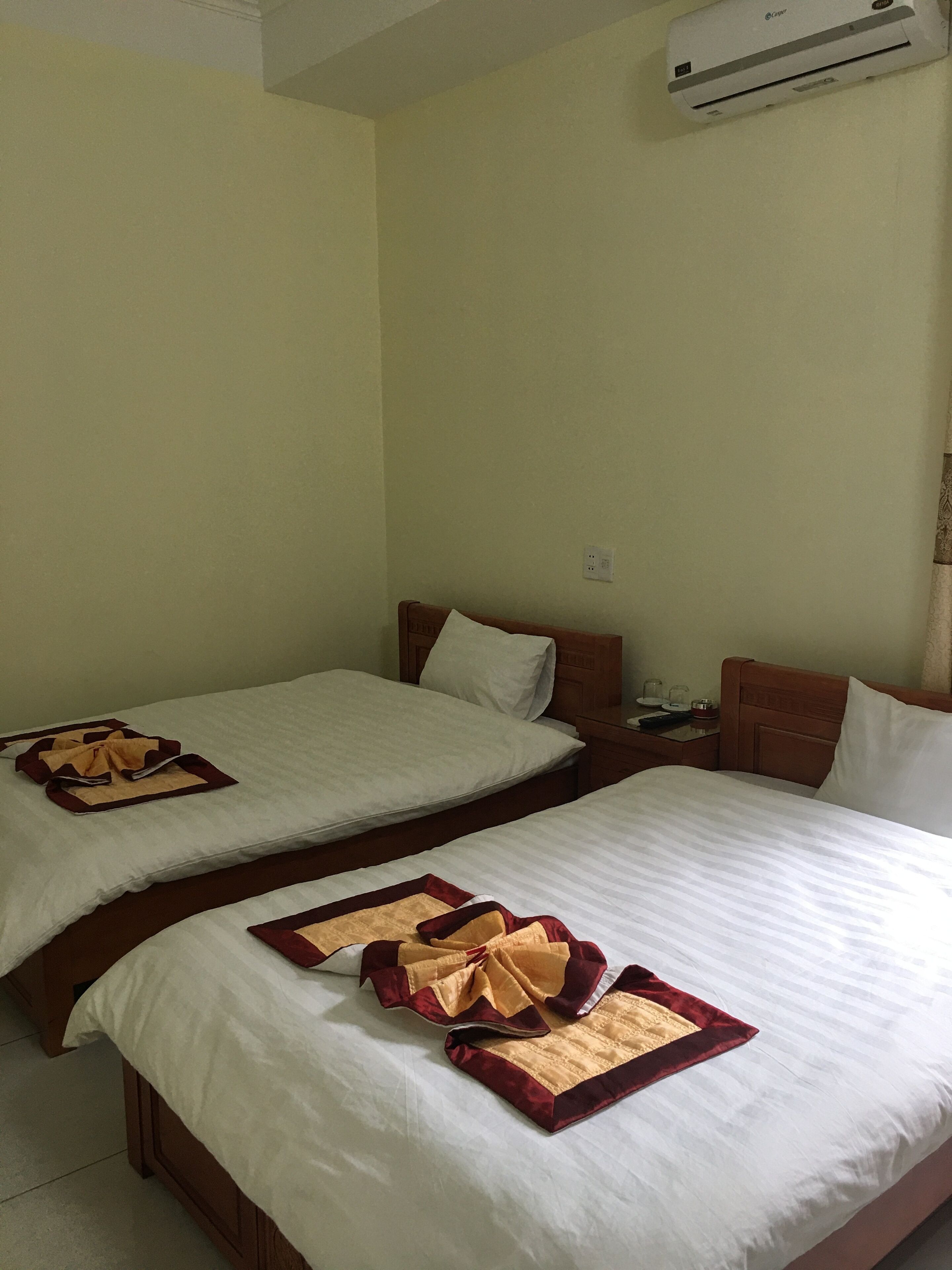Habitación de hotel con dos camas, cada una con cubrecama blanco y una manta decorativa roja y dorada. Cada cama tiene un cabecero de madera y una mesita de noche con una lámpara y una taza encima.