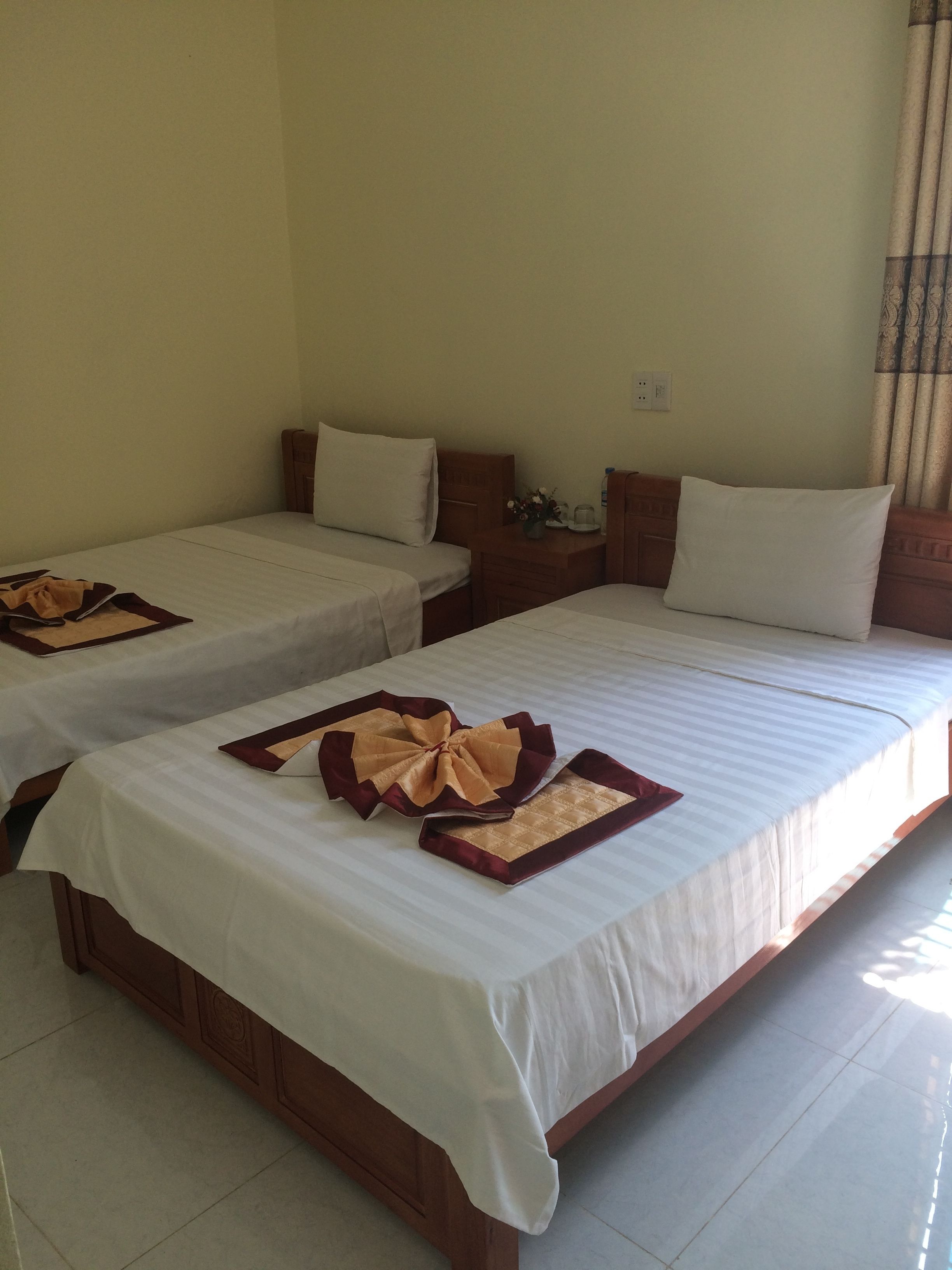 Two single beds with white linens and decorative pillows, a wooden headboard, a nightstand with a vase of flowers, and a tiled floor.