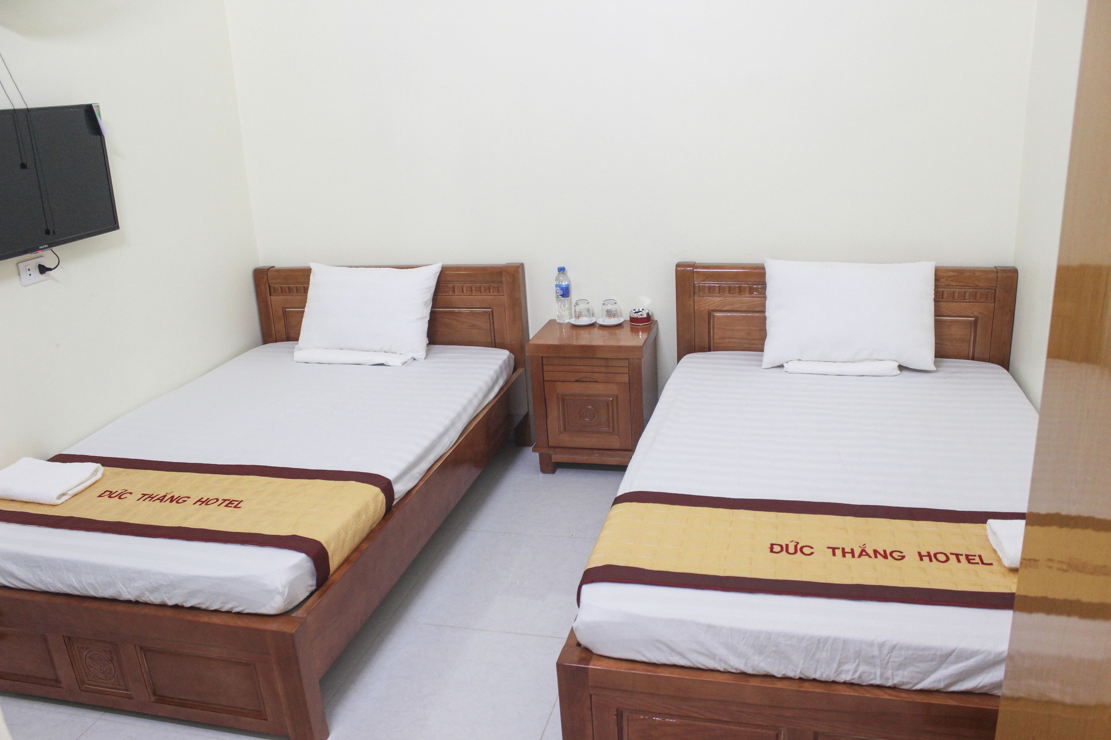 Een hotelkamer met twee eenpersoonsbedden, elk met een houten hoofdeinde en een beddeken met de tekst 'Duc Thanh Hotel'. Tussen de bedden staat een houten nachtkastje met een waterfles en glazen erop.
