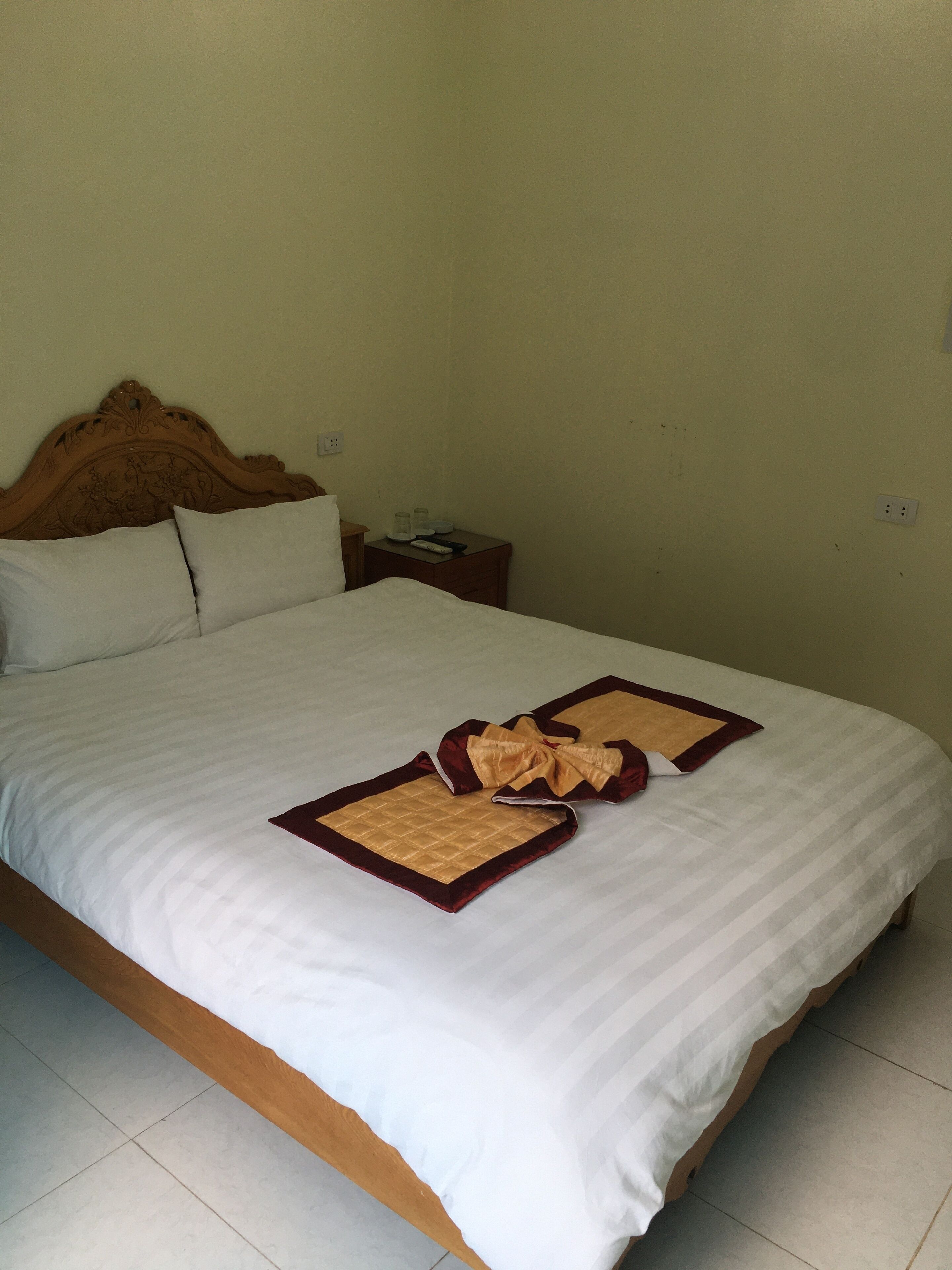 A neatly made bed with white bedding and two decorative pillows, a wooden headboard, a small bedside table with a lamp, and a power outlet on the wall.
