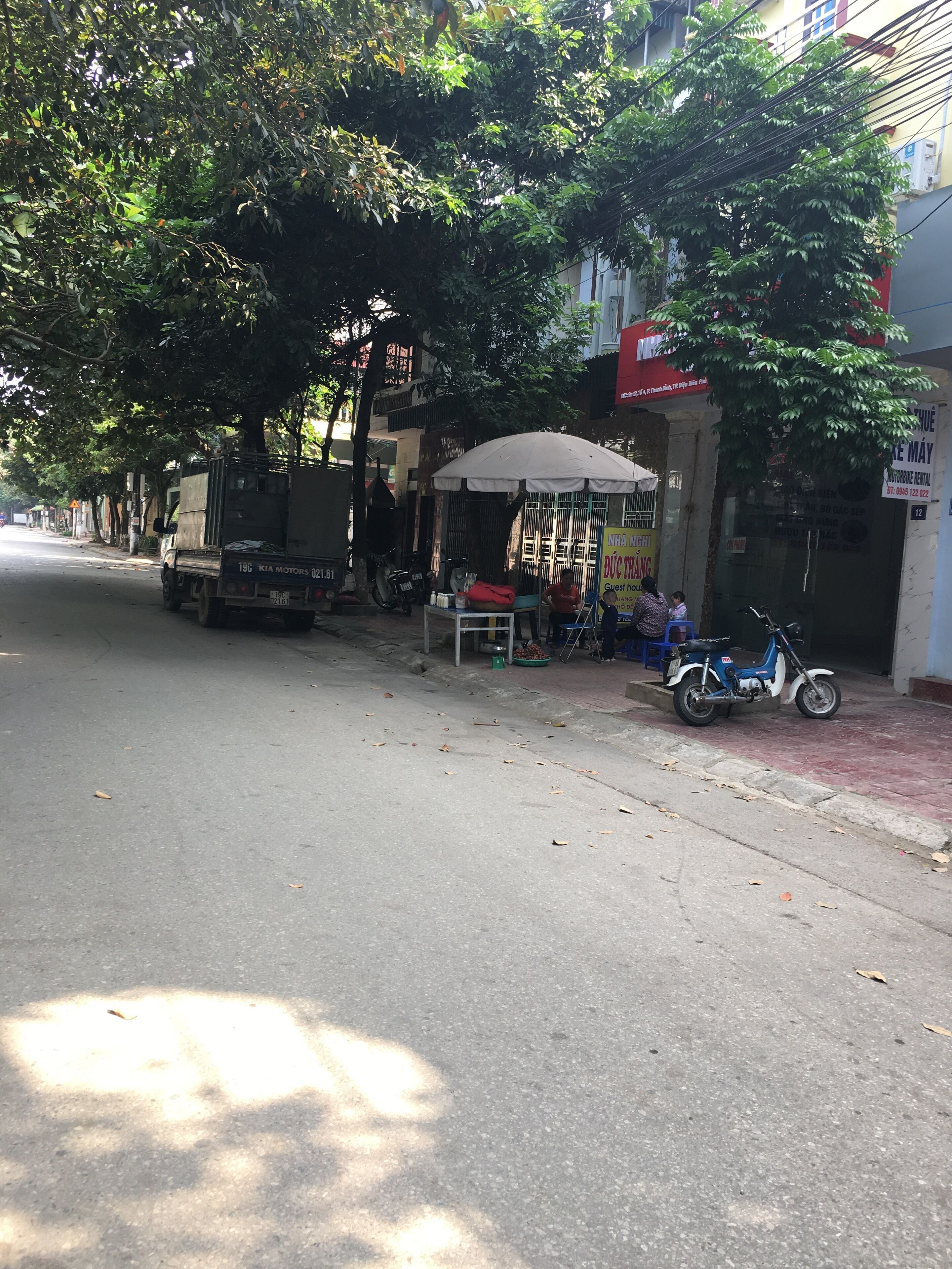 Una calle con un camión estacionado, un pequeño puesto de ventas bajo una tolda y una moto.