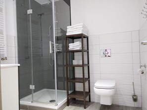 A modern bathroom with a glass-enclosed shower, a white toilet, and a wooden shelf with towels. - Hotel Babylon (Ringsheim)