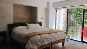 A modern bedroom with a large bed, a wooden headboard, and a view of a garden through a sliding glass door. - Lofts El Sueño (Puebla)