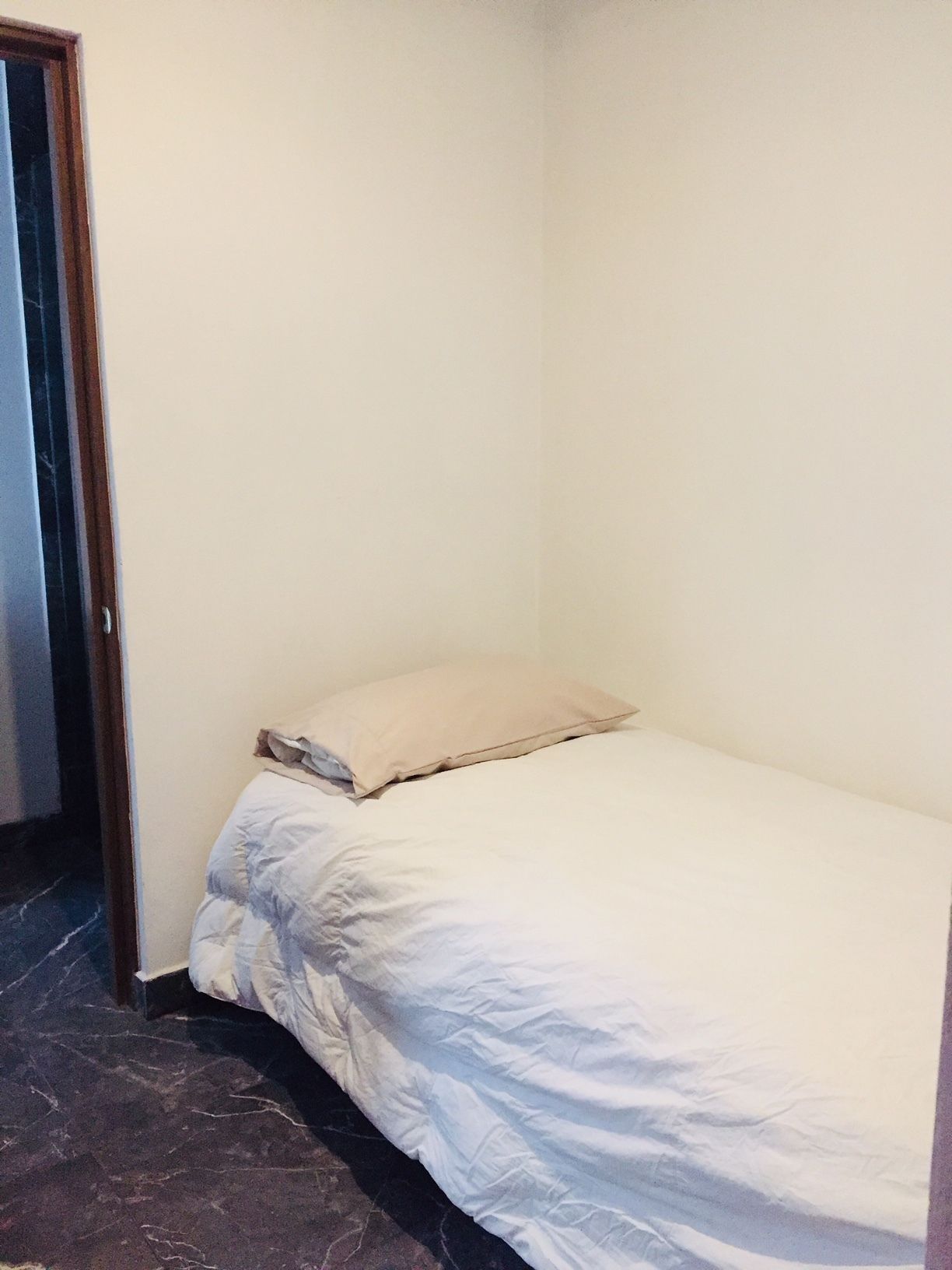 A small, unadorned room with a single bed, a beige pillow, and a plain white wall.