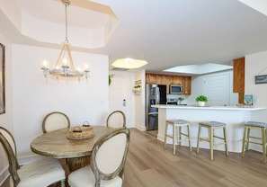 Un comedor con candelabro, mesa y sillas de madera, una cocina con armarios de madera y un refrigerador, y una zona de bar con taburetes.