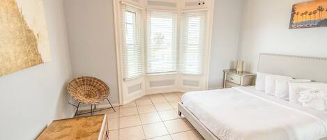 Un dormitorio con cama, ventilador de techo, ventana con persianas, una silla de mimbre y un cuadro en la pared.