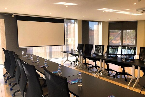 A conference room with multiple tables and chairs, a large screen, and windows with a view of buildings. - Mythos Boutique Hotel (Kigali)