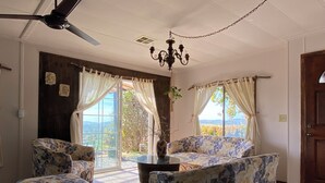 A room with a ceiling fan, a chandelier, a sofa, two armchairs, a coffee table, and a view through large windows. - Posada San Antonio (Valle de Guadalupe)