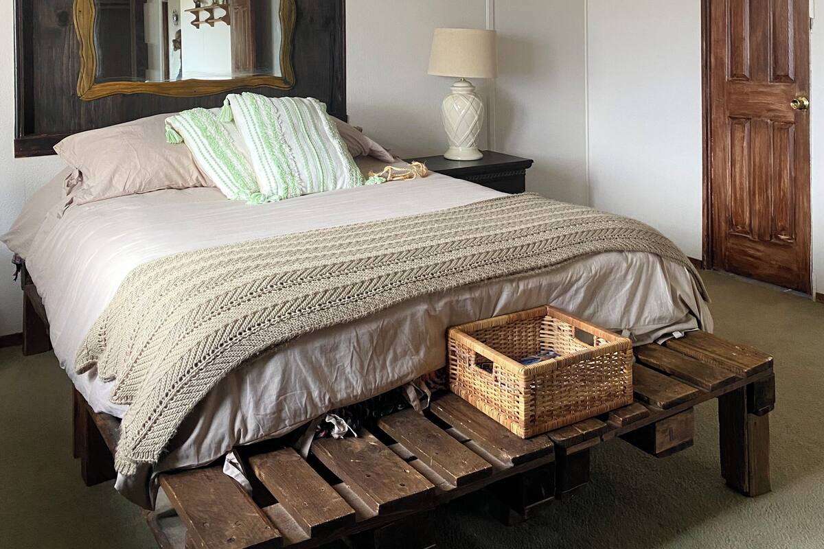 A bedroom with a bed, a ceiling fan, a mirror, a nightstand, a lamp, a wooden door, and a wooden bench.