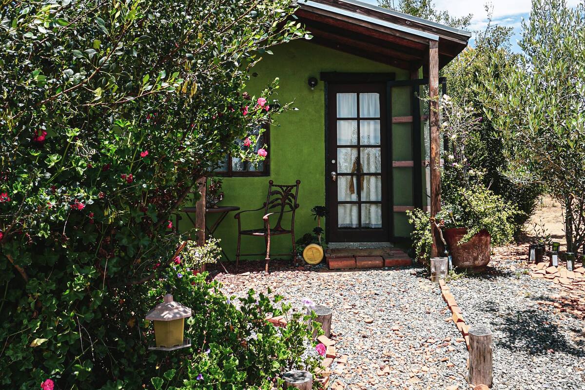 A small house with a green exterior, a wooden door, and a window. There is a garden with flowers and a gravel path leading to the door.