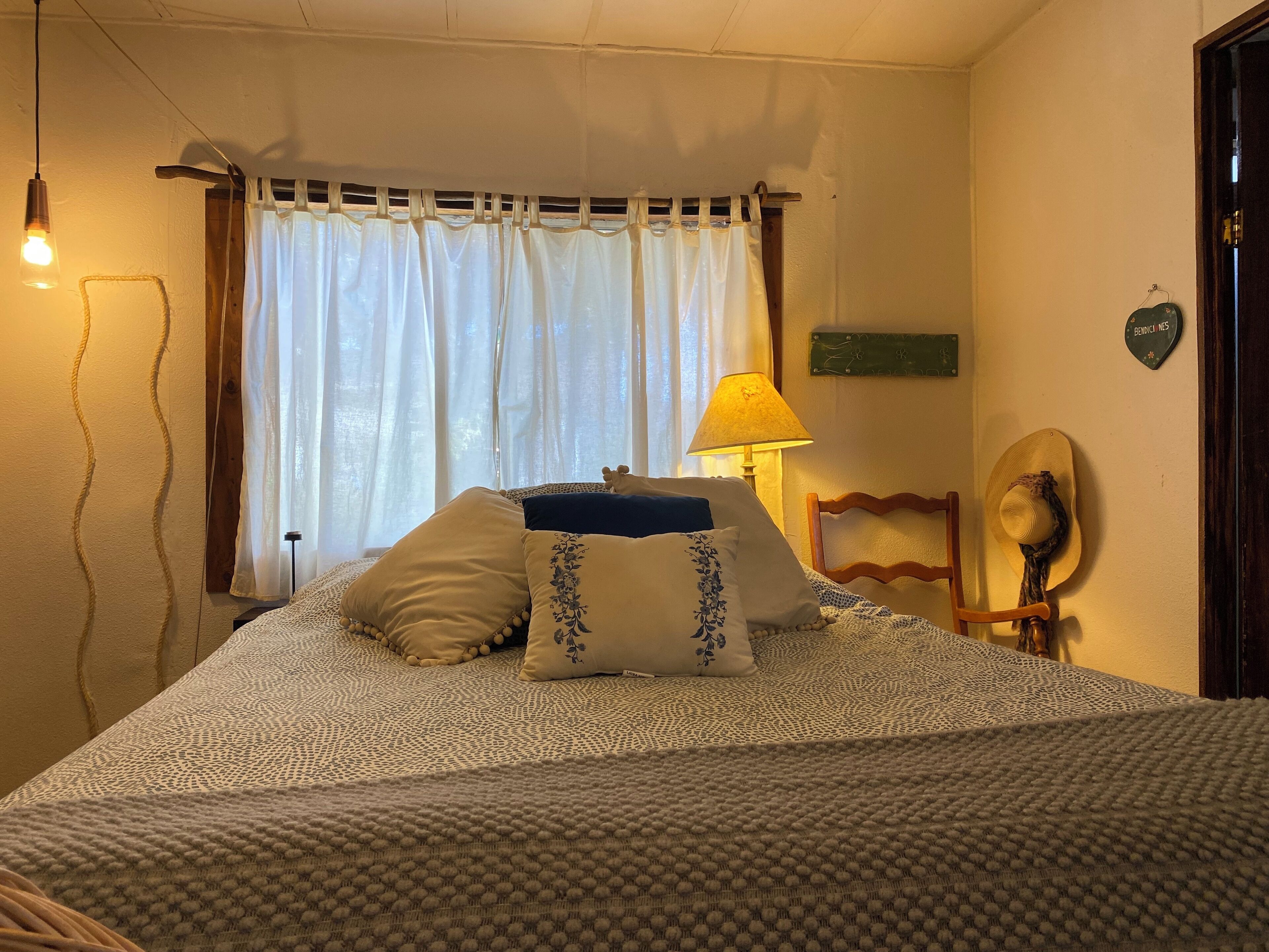 Un lit bien fait, avec une couvre-lit à motifs et des oreillers, une lampe sur une table de chevet et une fenêtre ornée de rideaux légers.