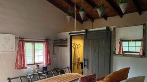 A bedroom with a bed, pillows, a window with curtains, a wooden door, and a hanging plant. - Posada San Antonio (Valle de Guadalupe)