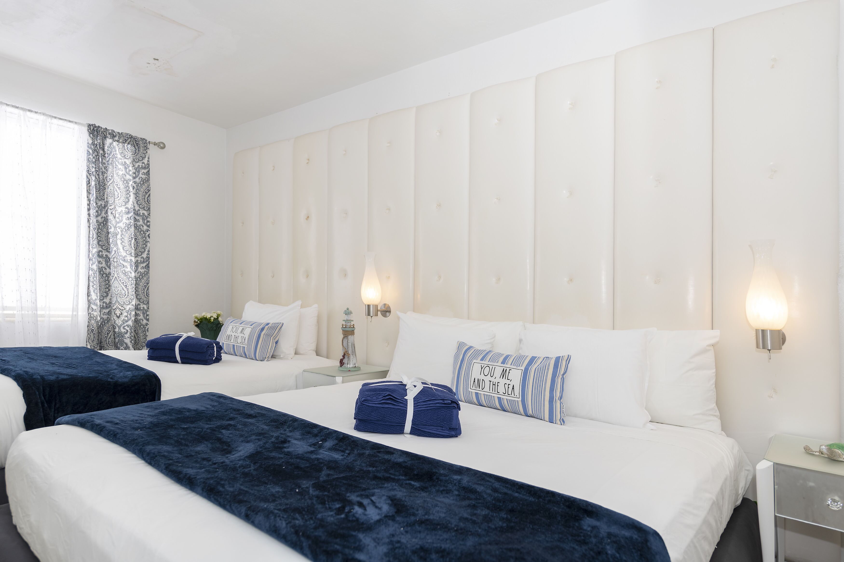 A hotel room with two beds, a large window with curtains, a headboard with lights, and a bedside table with a lamp.