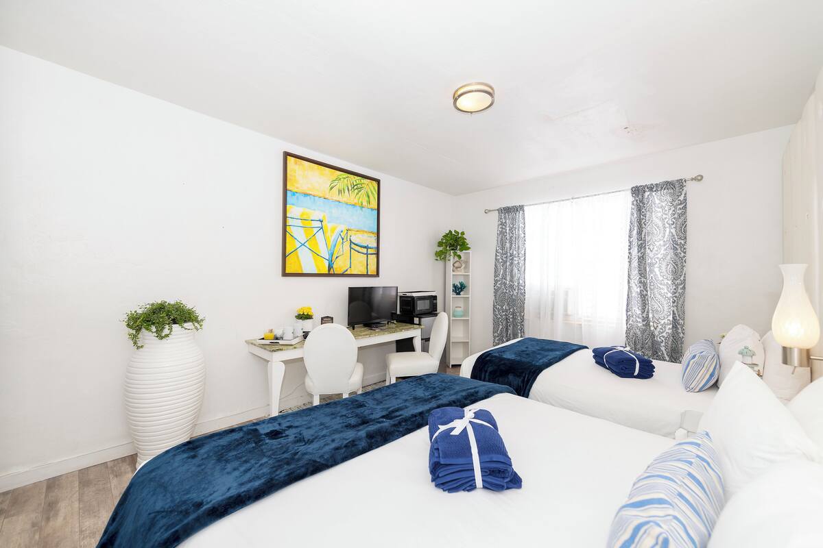A hotel room with two beds, a desk with a computer, a TV, and a large window with curtains.