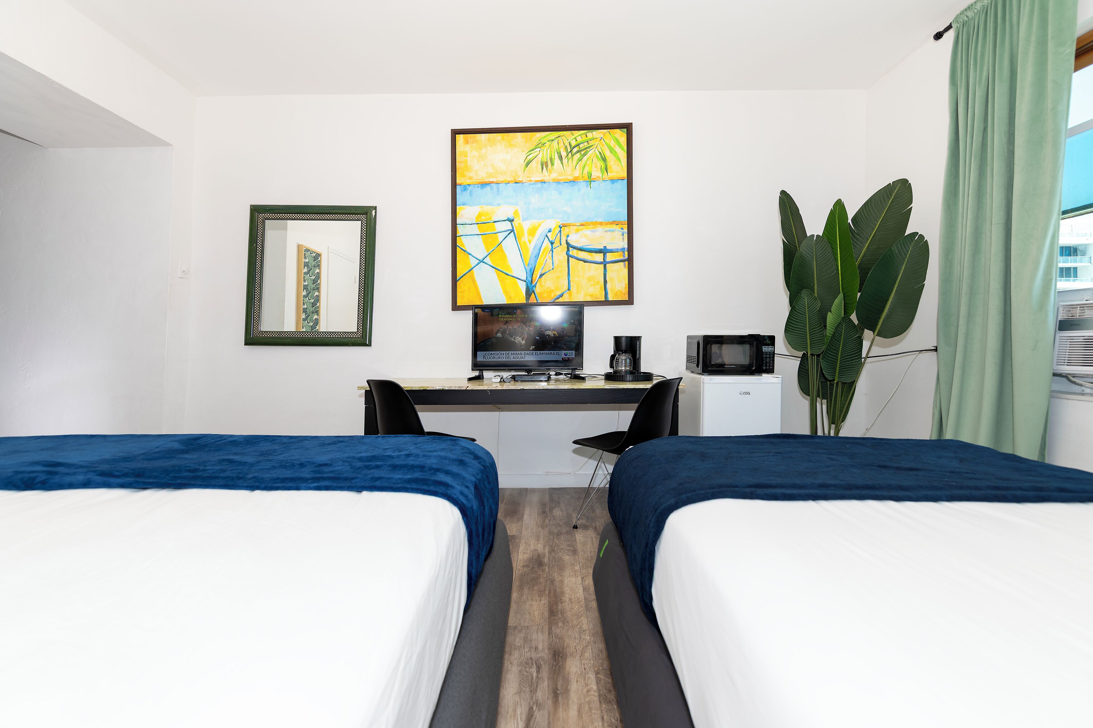 Habitación de hotel con dos camas, un escritorio con computadora, televisor, cafetera y una planta grande.