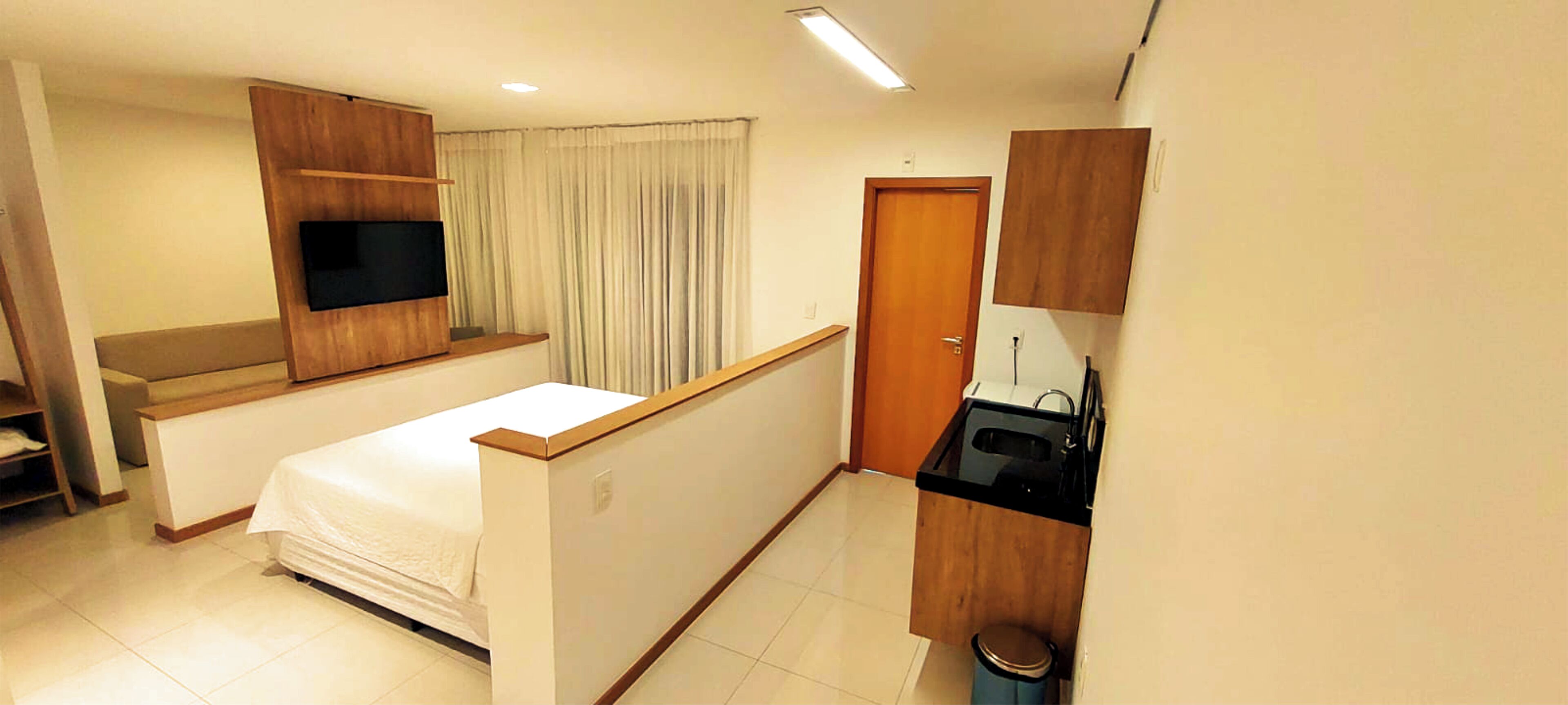 A compact hotel room with a bed, a small desk, a TV mounted on the wall, a kitchenette, and a door.