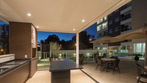 Un espace cuisine extérieur moderne, doté d’un îlot central, de sièges et offrant une vue sur l’intérieur du bâtiment.