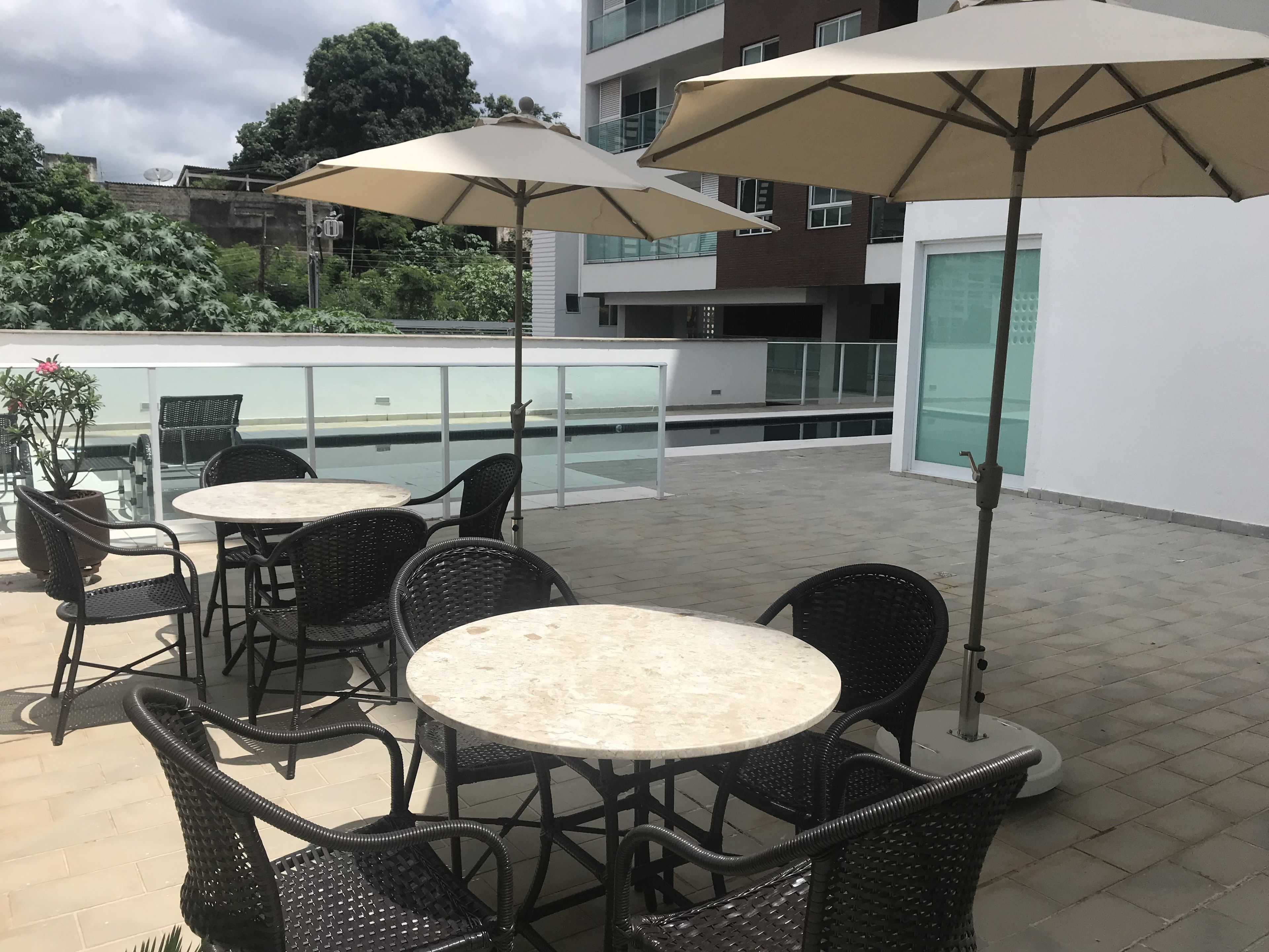 An outdoor patio area with round tables and chairs, large umbrellas, and a view of a swimming pool and buildings in the background.