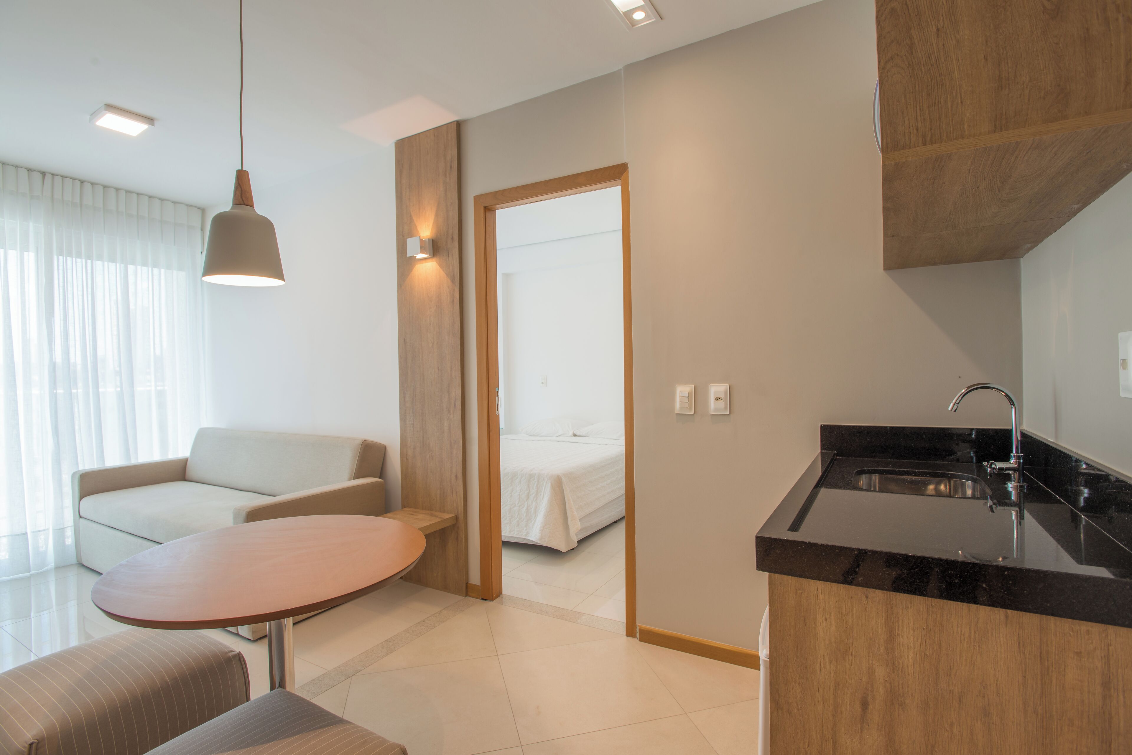 A modern hotel room with a kitchenette, a dining area with a table and chairs, a sofa, and a bed in the background.
