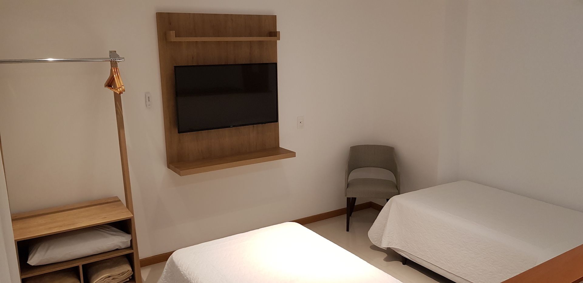 A small, clean room with a bed, a chair, a shelf with folded towels, and a mounted TV.