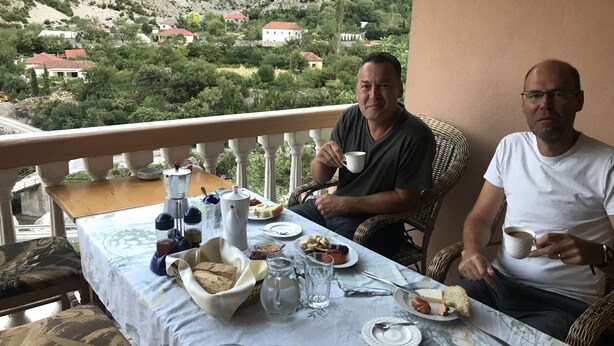 Zwei Männer essen an einem Tisch mit Blick auf eine Berglandschaft, weißer Tischdecke und verschiedenen Speisen.