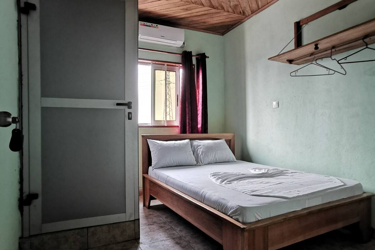 A bedroom with a wooden ceiling, a bed with a white mattress, and a window with curtains.