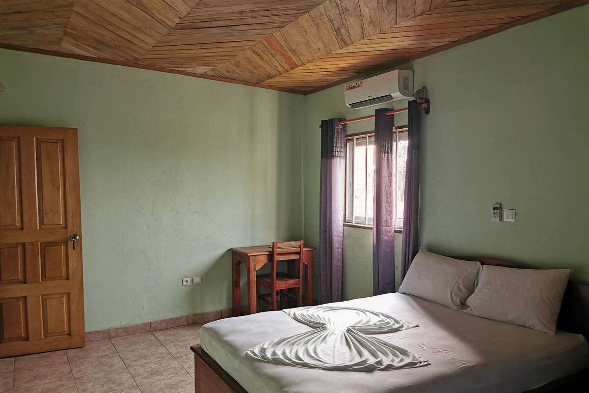 A bedroom with a wooden ceiling, a bed with a white bedspread, a wooden nightstand, a window with curtains, and a door.