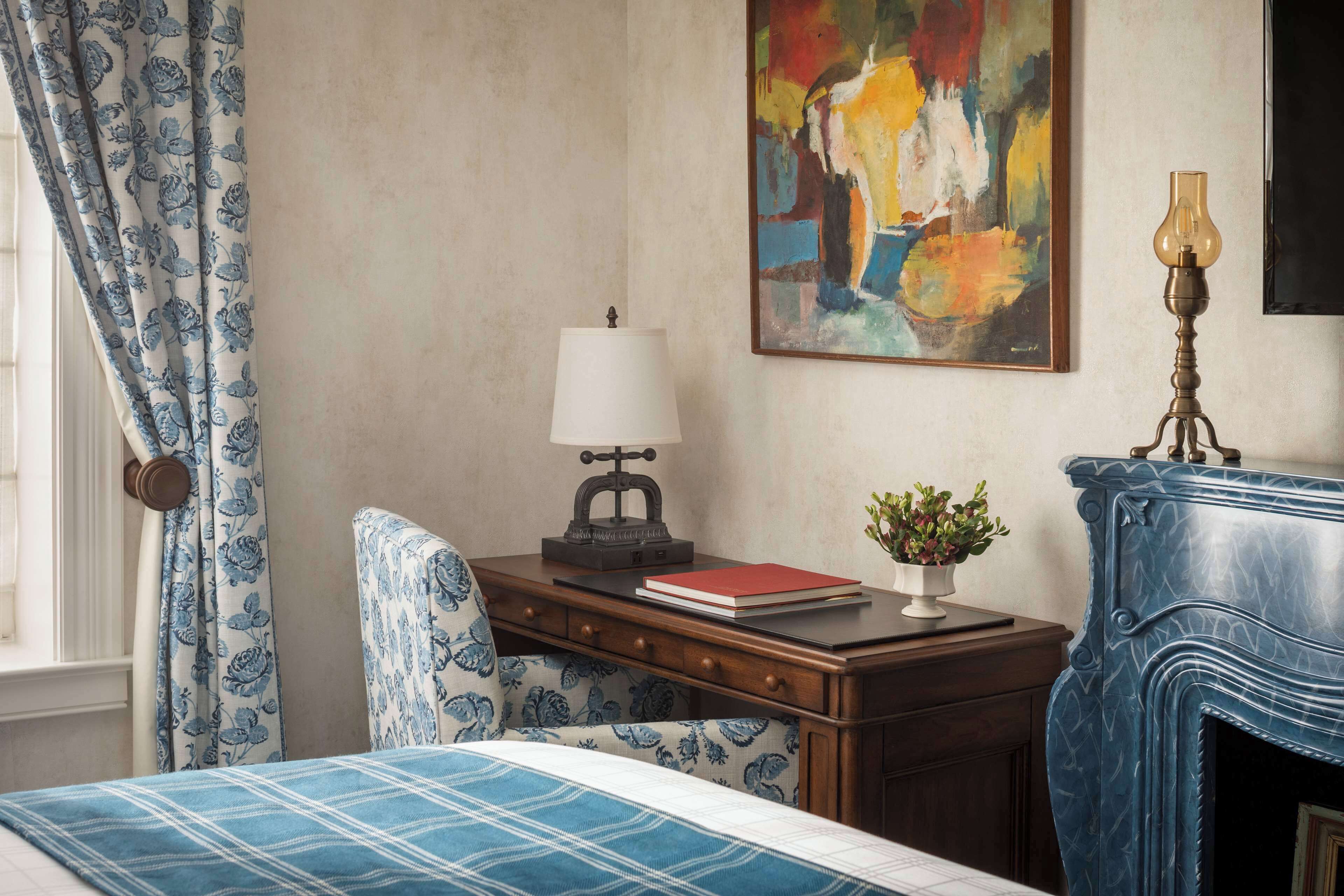 Quarto com cama, escrivaninha com luminária, cadeira, quadro e lareira.