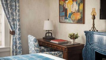 Quarto com cama, escrivaninha com luminária, cadeira, quadro e lareira.