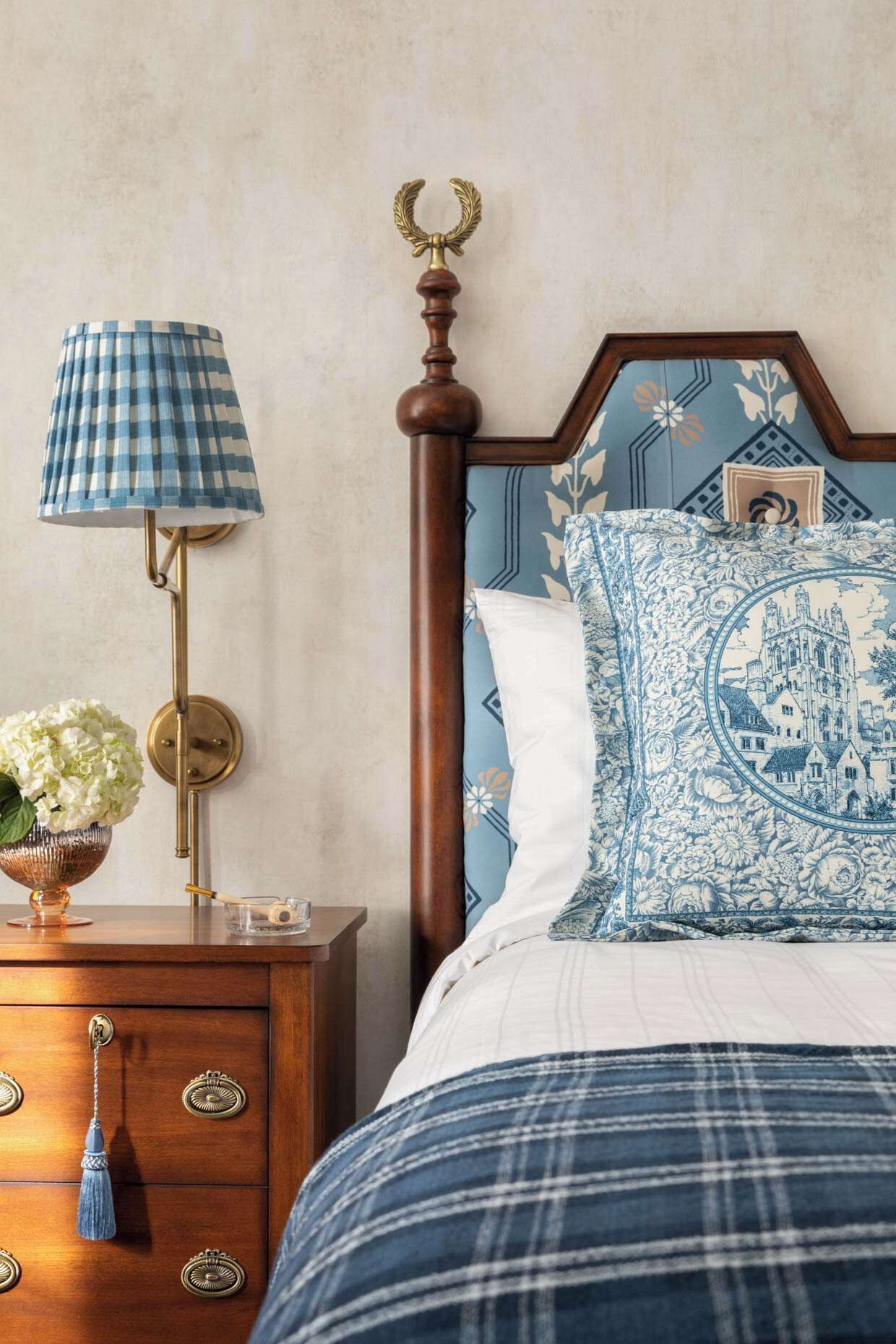 A bedroom with a wooden headboard, a blue and white patterned bedspread, a matching pillow, a bedside table with a lamp, and a vase with flowers.