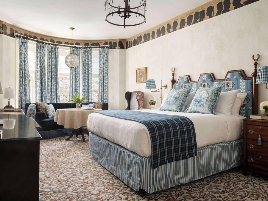 A bedroom with a large bed, a desk, and a chair. There is a window with curtains, a chandelier, and a wall with a patterned design.