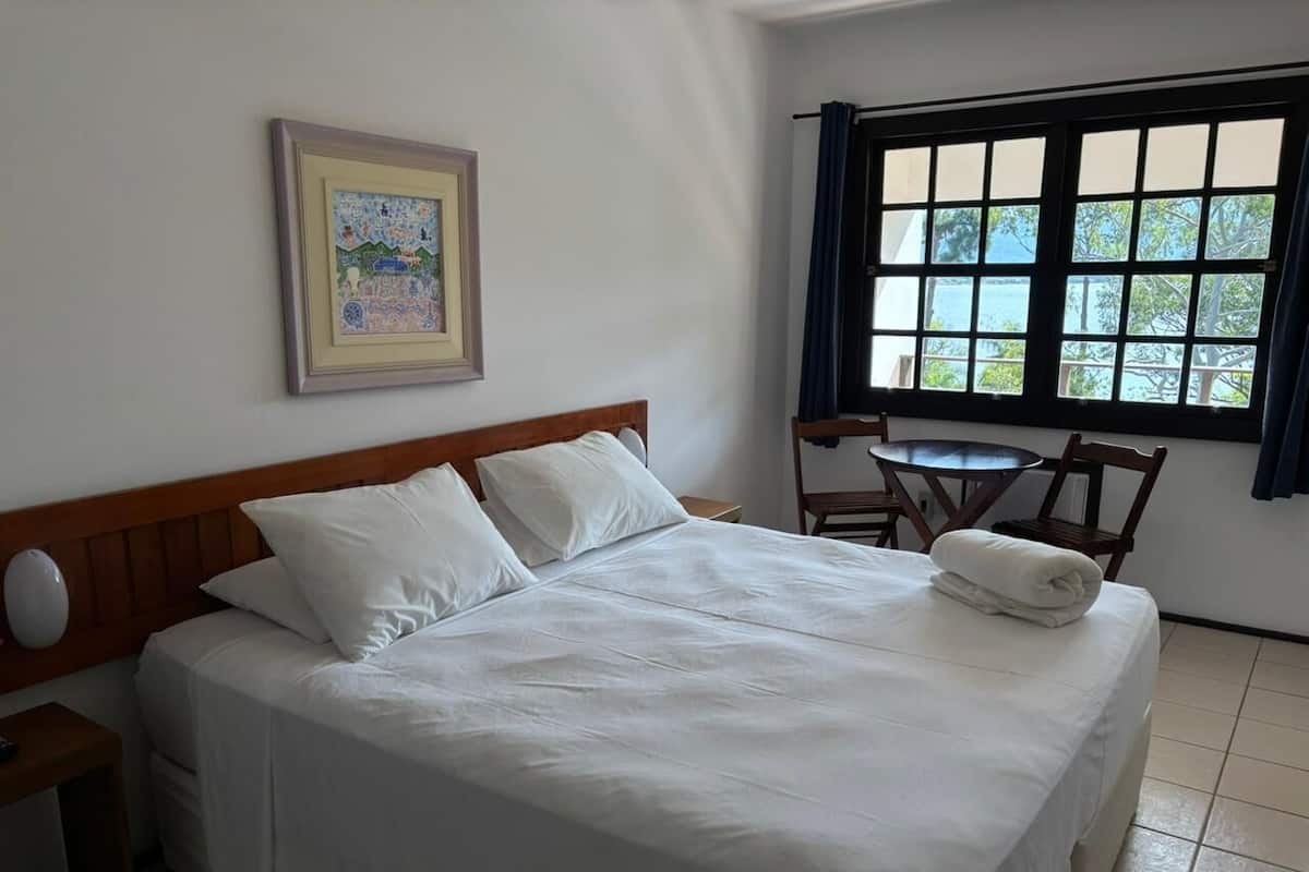 Un lit bien fait, avec du linge de lit blanc, une tête de lit en bois, une petite table de chevet, un tableau encadré au mur et une fenêtre avec des rideaux.