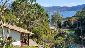 A house with a patio, surrounded by lush greenery and a view of a lake and distant mountains. - Residencial Cabanas da Praia Mole (Florianópolis)