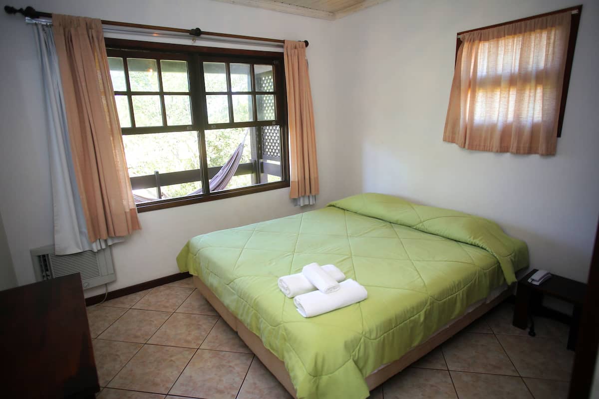 Une chambre avec un lit simple, une couverture verte, une fenêtre avec des rideaux, un radiateur et une petite table avec une chaise.