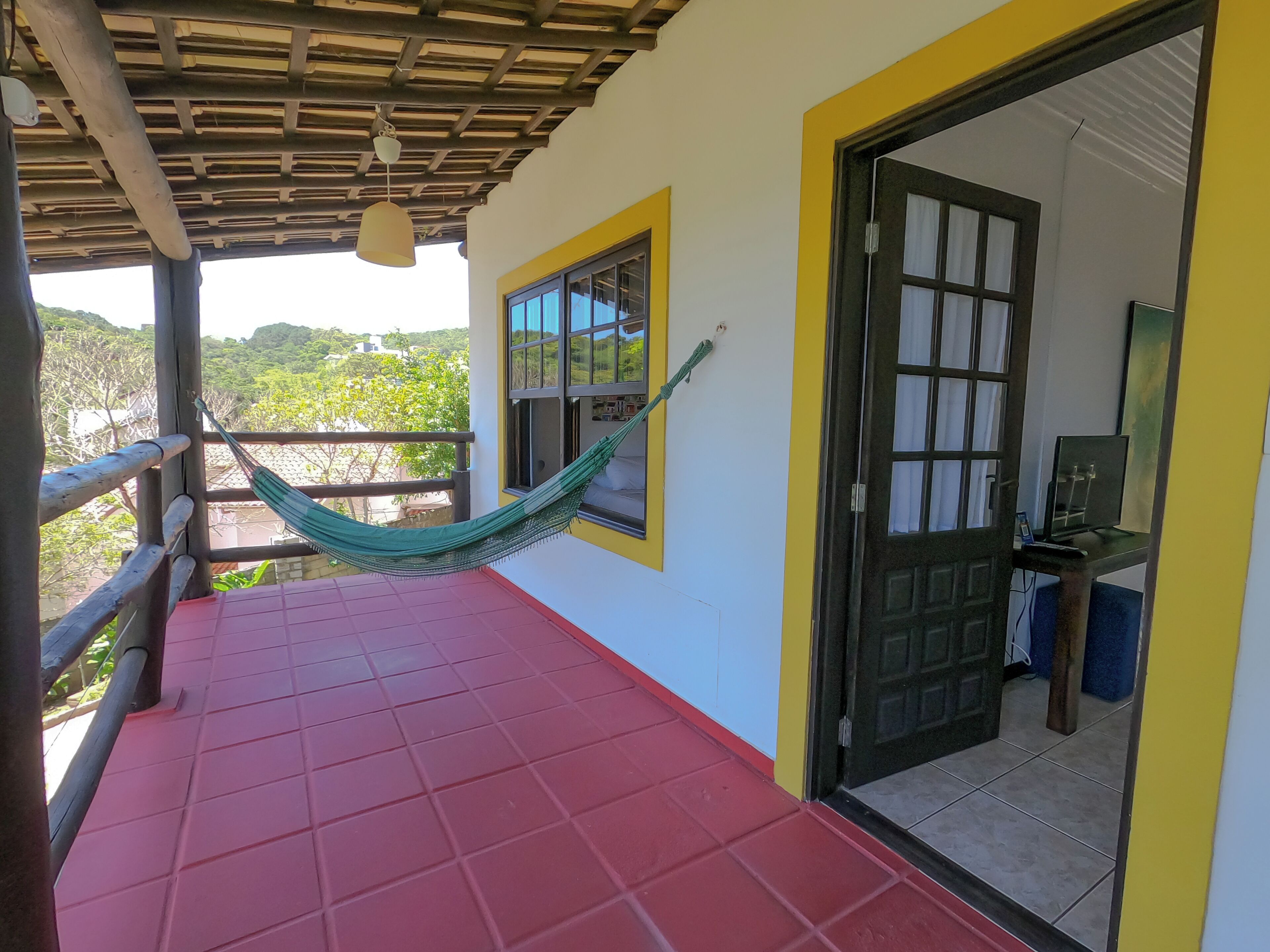 Un balcon avec un sol rouge, un hamac, une rambarde en bois et une porte donnant sur une pièce dont le sol est carrelé.