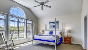 A bedroom with a large window, a bed, a ceiling fan, and a nightstand.