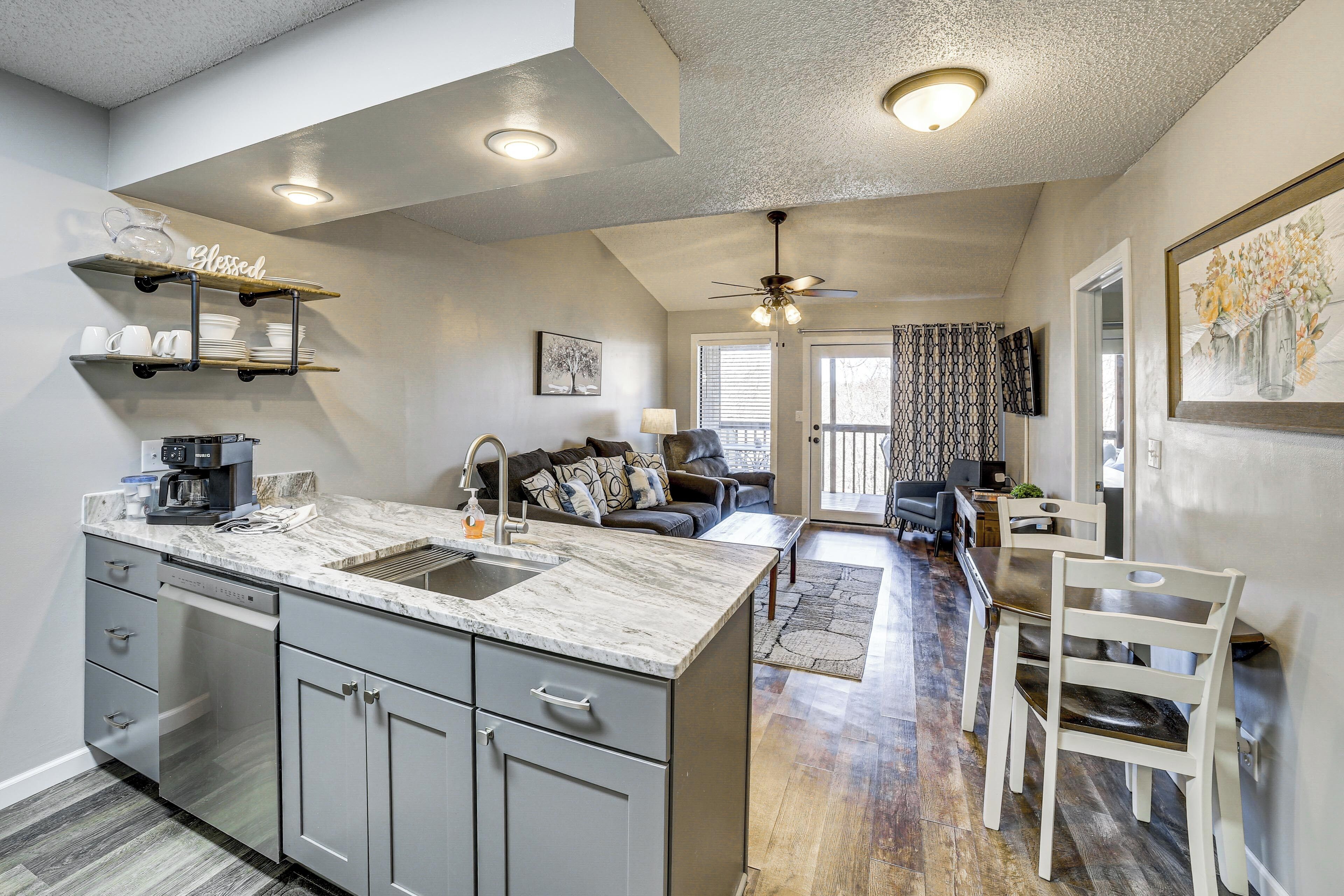 A modern kitchen with a marble countertop, grey cabinets, and a built-in dishwasher. There is a dining area with a table and chairs, and a living room with a sofa and a ceiling fan.